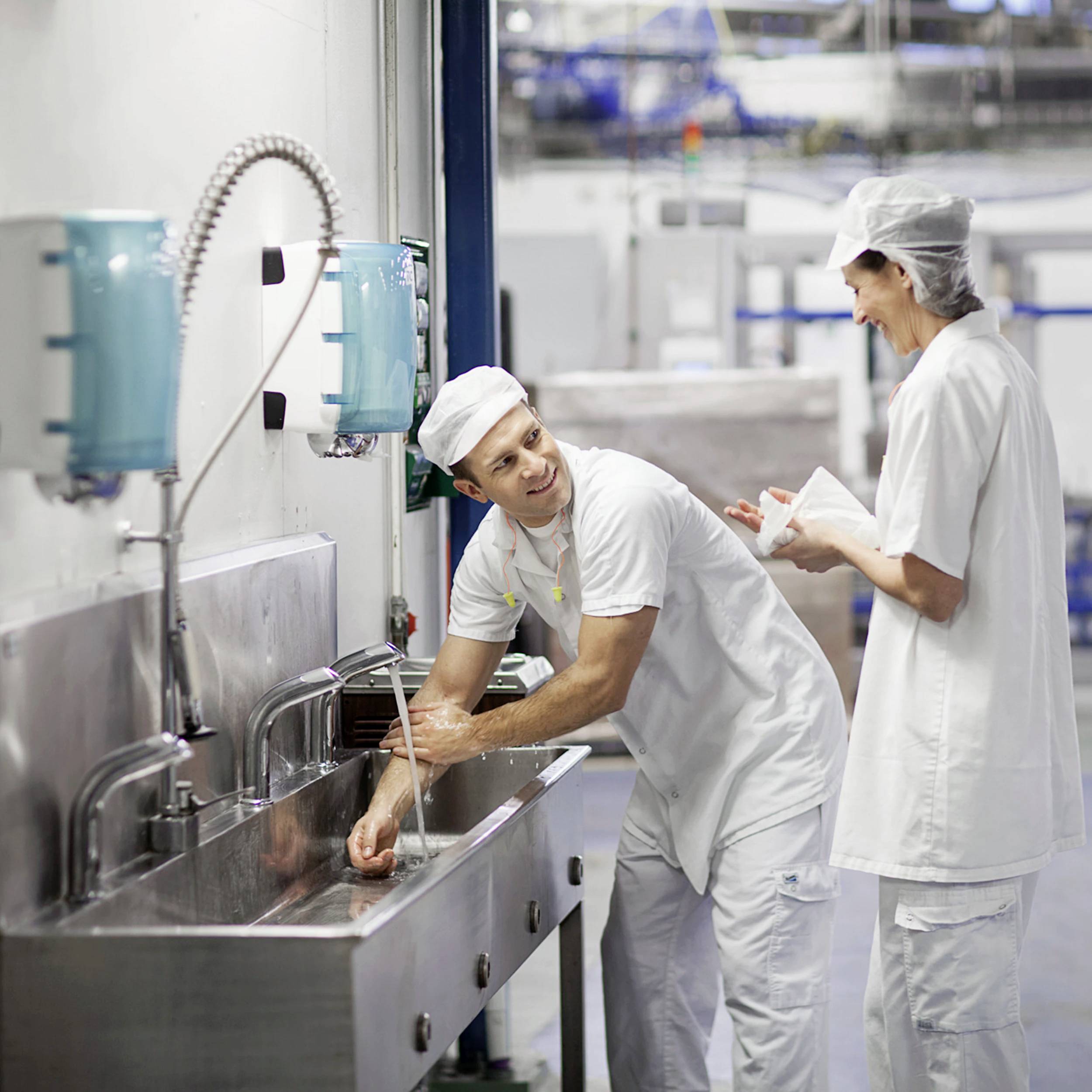 Deux personnes en tenue de travail se lavent les mains à un lavabo industriel. La pièce ressemble à une usine ou à une grande cuisine.