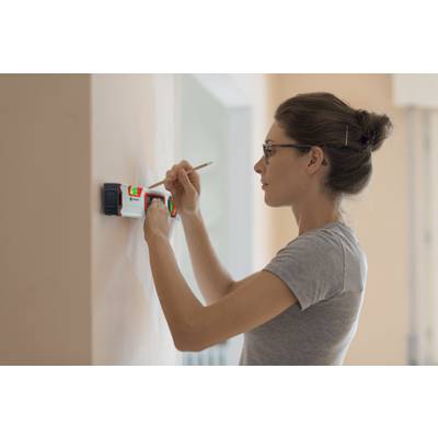 Une femme utilise un appareil de mesure électronique sur un mur, tout en faisant une marque avec un stylo. Elle porte un t-shirt gris.