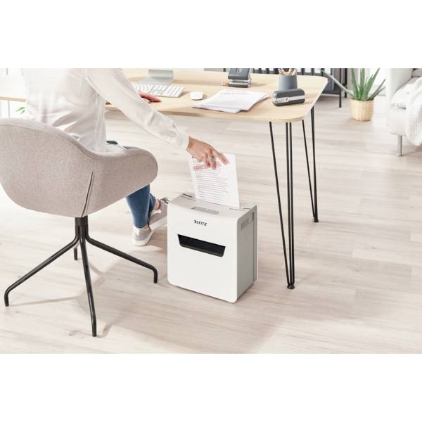 Une personne est assise à un bureau et utilise un destructeur de documents pour déchiqueter une feuille de papier. Des meubles de bureau sont visibles en arrière-plan.