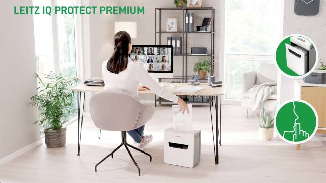 Une femme est assise à son bureau et utilise une déchiqueteuse. Un appel vidéo est visible sur l'écran. Texte : 'LEITZ IQ PROTECT PREMIUM'.