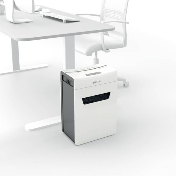 Un destructeur de documents se trouve à côté d'un bureau avec une chaise de bureau. La pièce a un aspect moderne et minimaliste.