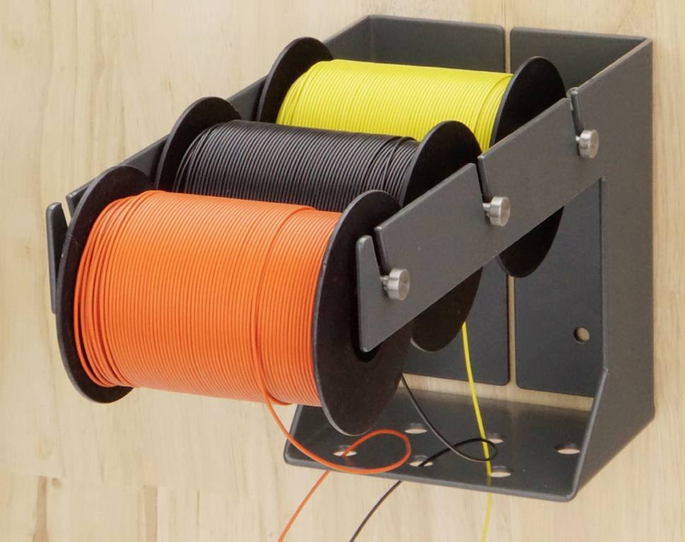 Trois bobines de fil en orange, noir et jaune sont fixées sur un support métallique contre un mur en bois.