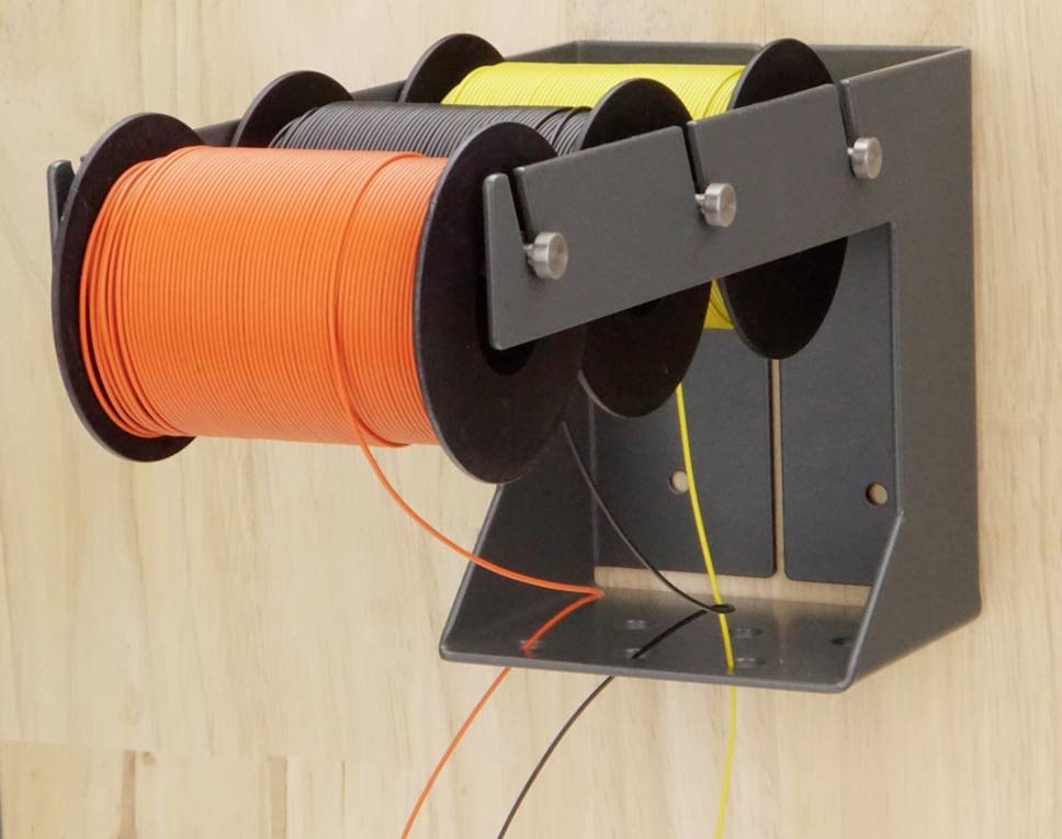 Trois bobines de fils colorés en orange, noir et jaune, montées sur un support fixé au mur, devant un fond en bois.