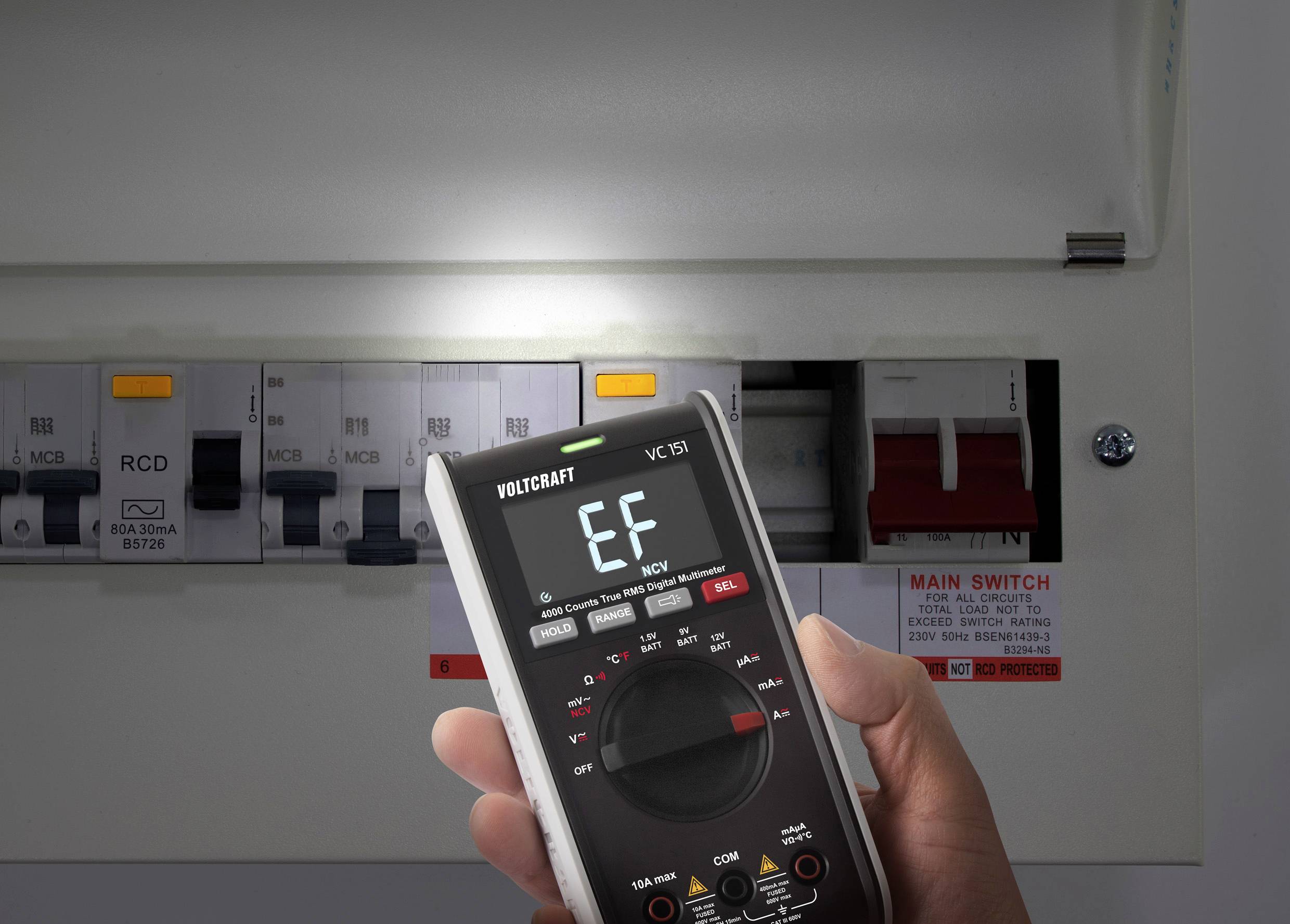 Une main tient un multimètre numérique affichant 'EF' devant un armoire électrique ouverte avec des disjoncteurs et un interrupteur principal.