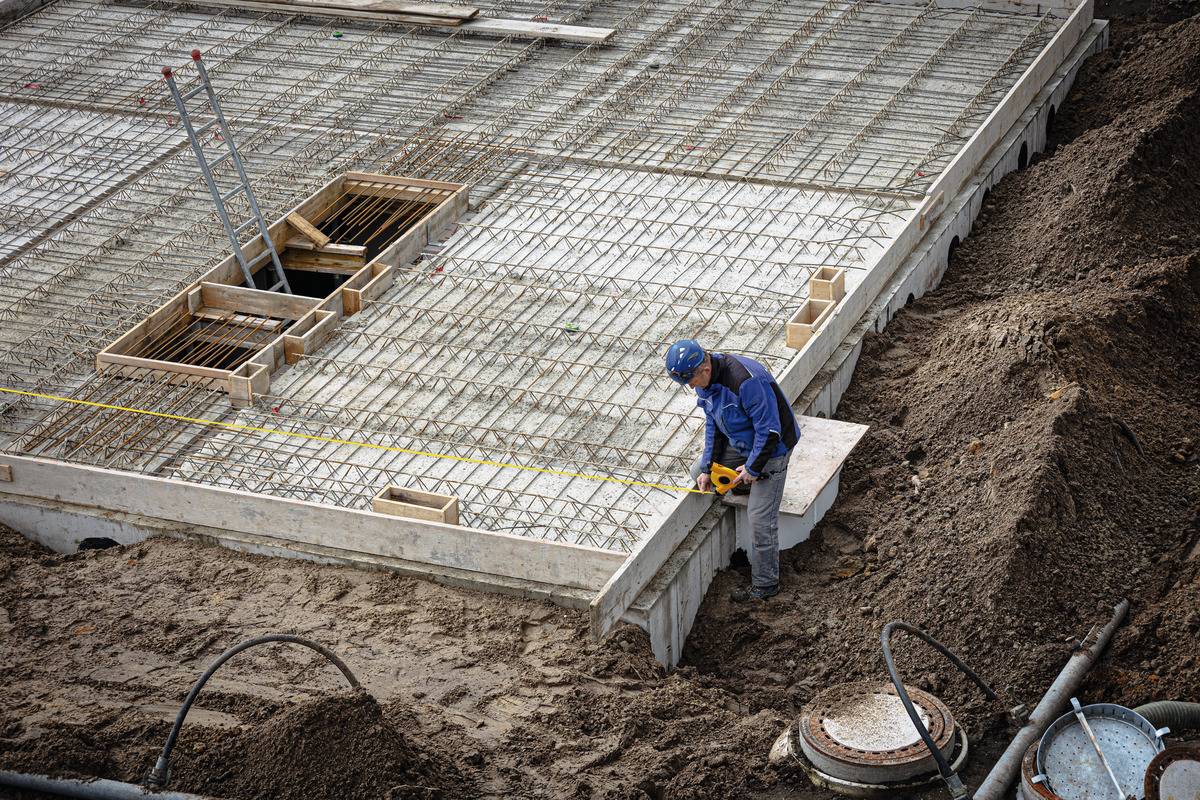 Un ouvrier mesure une dalle en béton armé sur un chantier. Grand espace ouvert, béton et acier apparents. Terre et outils de construction au premier plan.