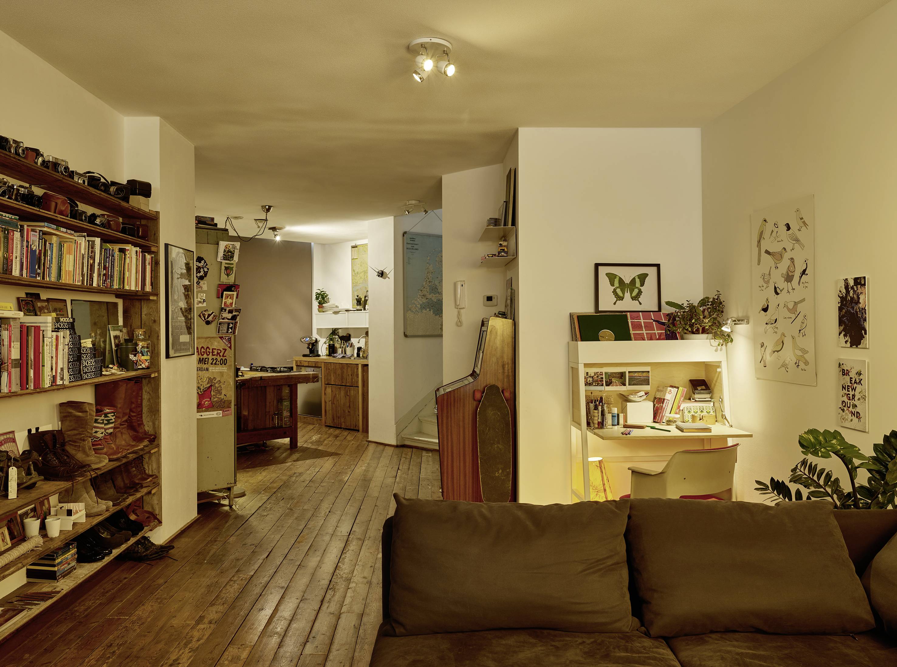 Salon avec sol en bois, bibliothèque, petit bureau et plantes. Cuisine ouverte en arrière-plan. Éclairage chaleureux et accueillant.