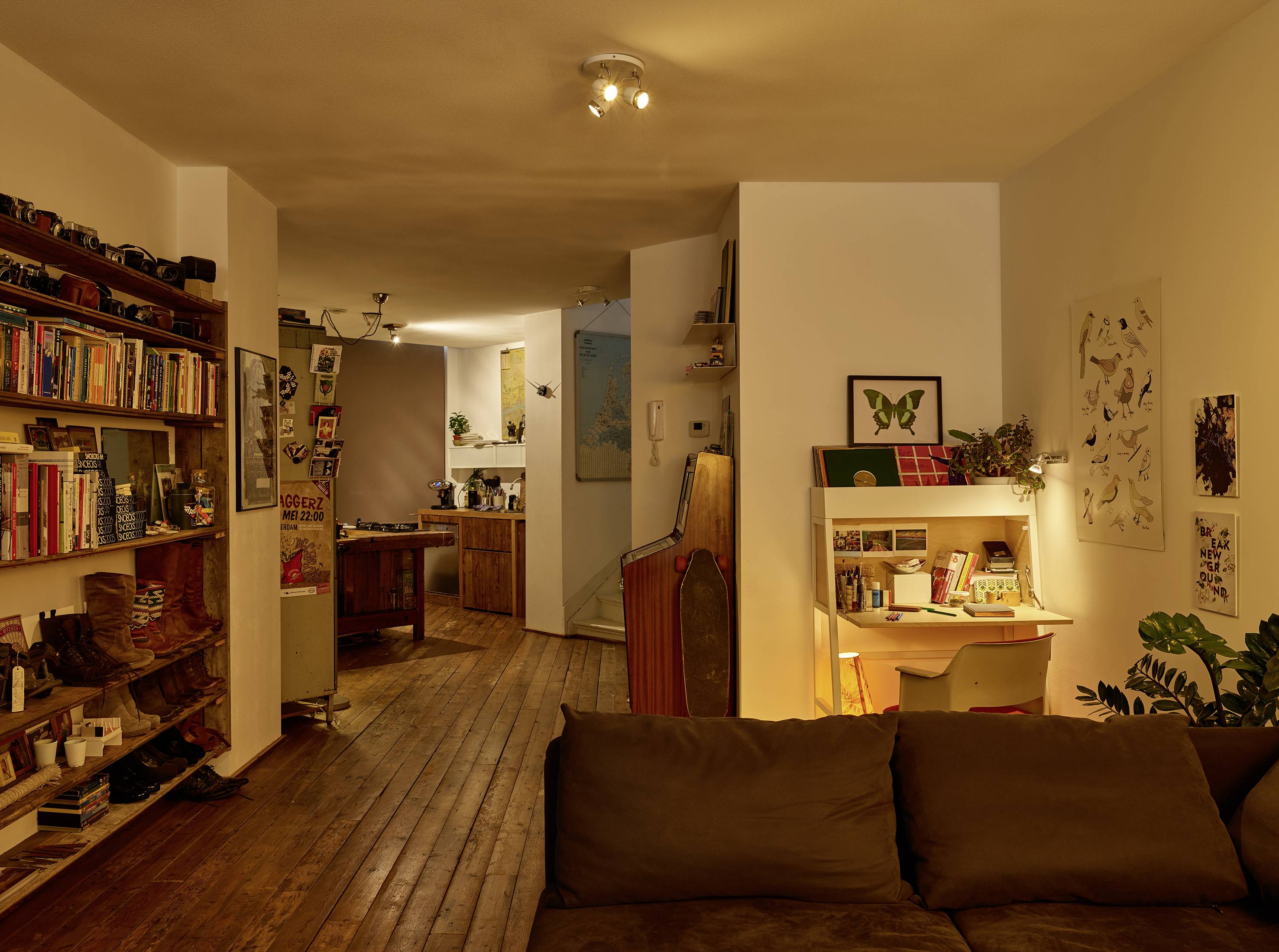 Salon confortable avec une bibliothèque, un canapé et un bureau. À l'arrière-plan, une cuisine ouverte baignée de lumière chaleureuse.