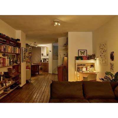 Salon confortable avec une bibliothèque, un canapé et un bureau. À l'arrière-plan, une cuisine ouverte baignée de lumière chaleureuse.