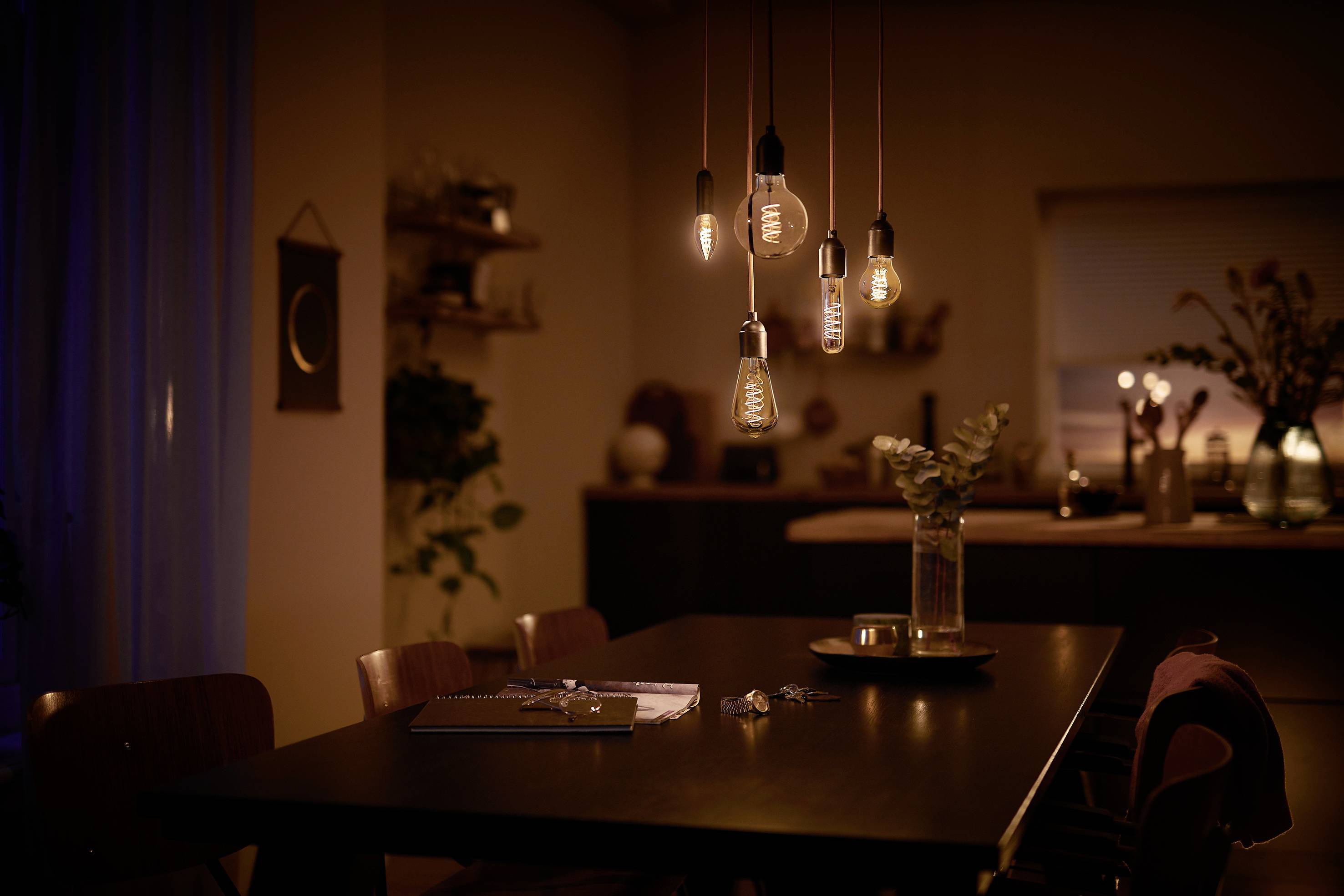 Une salle à manger chaleureusement éclairée avec une table sur laquelle sont posés un carnet et un vase. Plusieurs ampoules pendent du plafond.