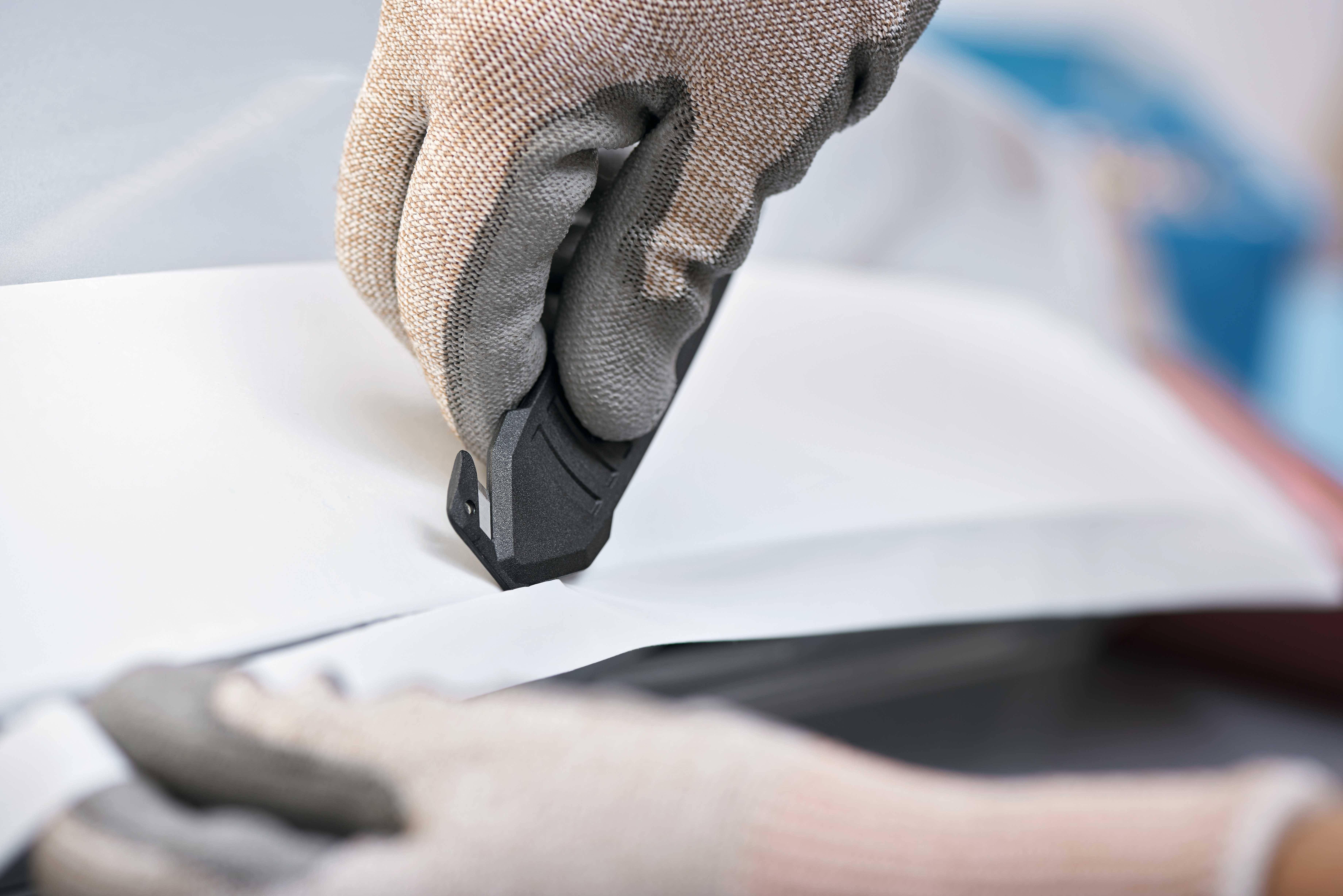 Une personne portant des gants coupe du papier épais ou du carton avec un cutter sur une surface lisse.