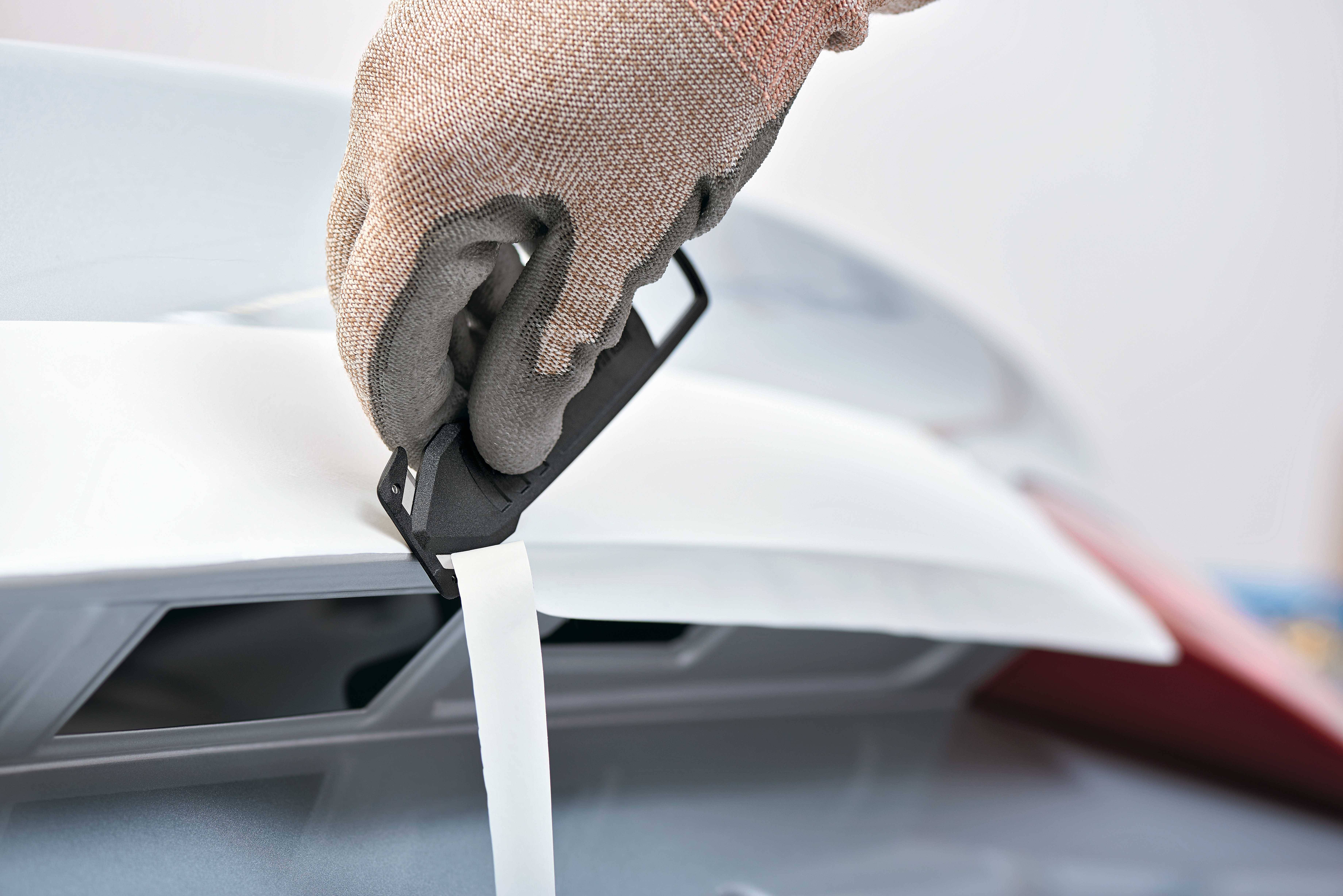 Une main portant un gant de travail utilise un outil de coupe pour détacher un ruban adhésif d'une surface.