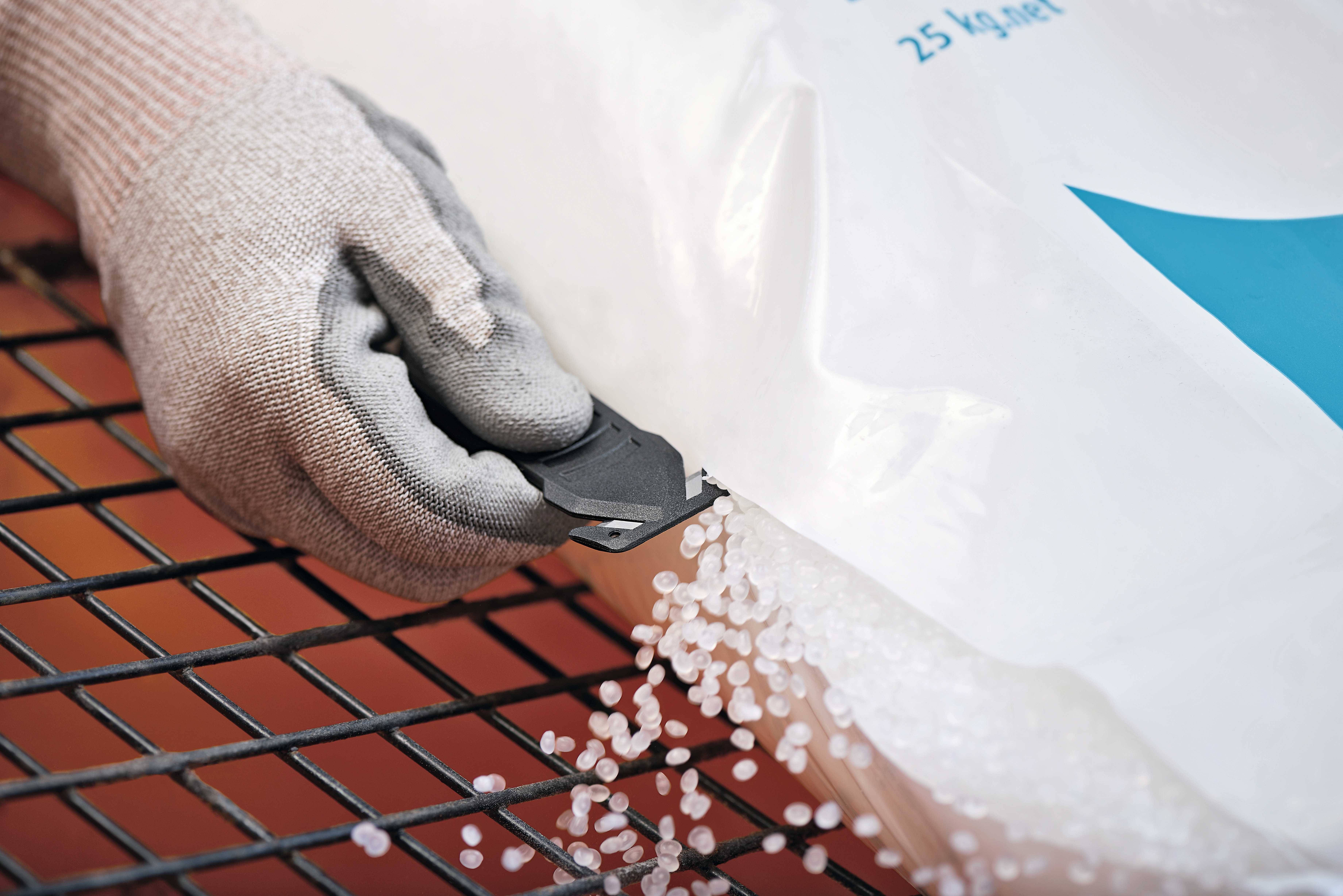 Une main gantée ouvre un sac à l'aide d'un couteau, et des granulés de plastique blanc s'écoulent sur une grille métallique.