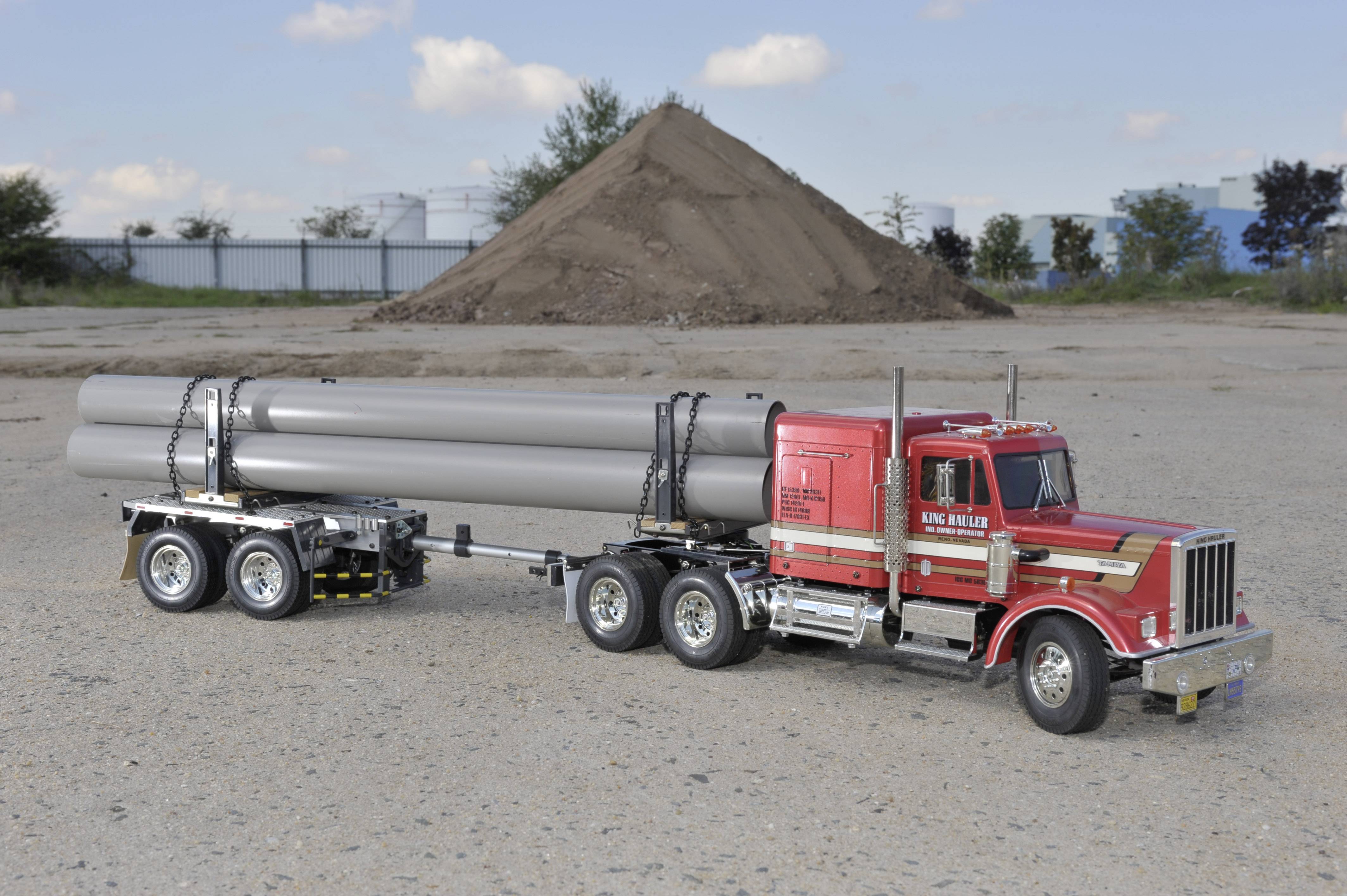 Un camion rouge avec une remorque transporte plusieurs longs tubes sur un chantier de construction, avec une colline de terre en arrière-plan.