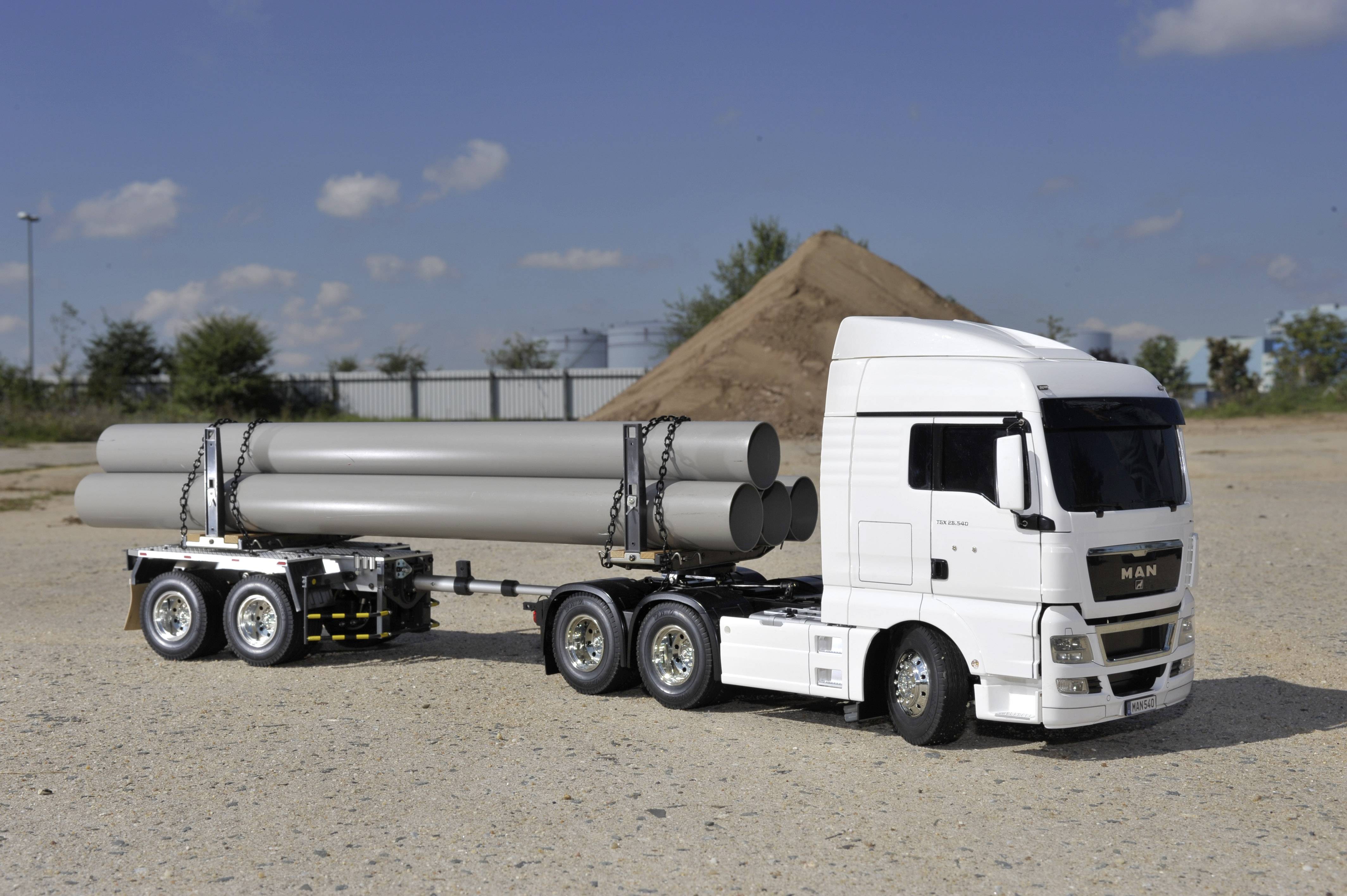 Un camion blanc transporte des tuyaux gris sur une remorque, stationné sur un chantier de construction avec un tas de terre en arrière-plan.
