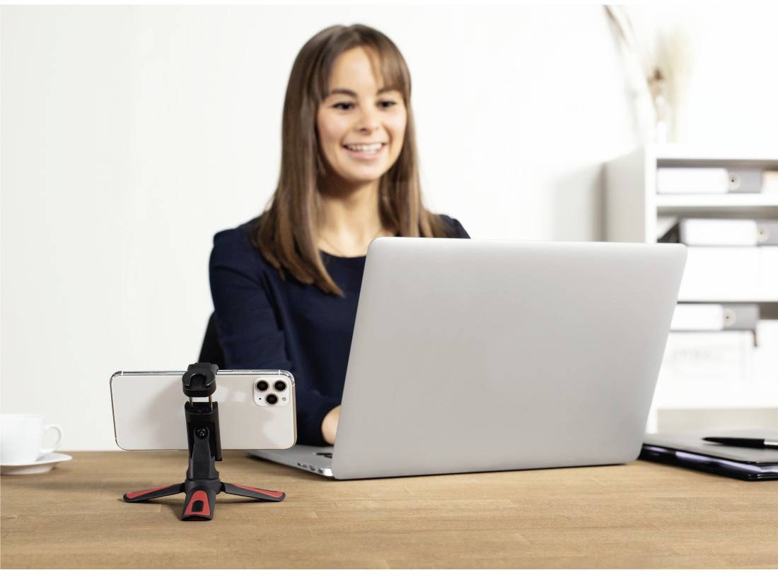 Une femme est assise à une table avec un ordinateur portable et un smartphone sur support. Elle sourit en regardant l'écran. Arrière-plan de bureau.