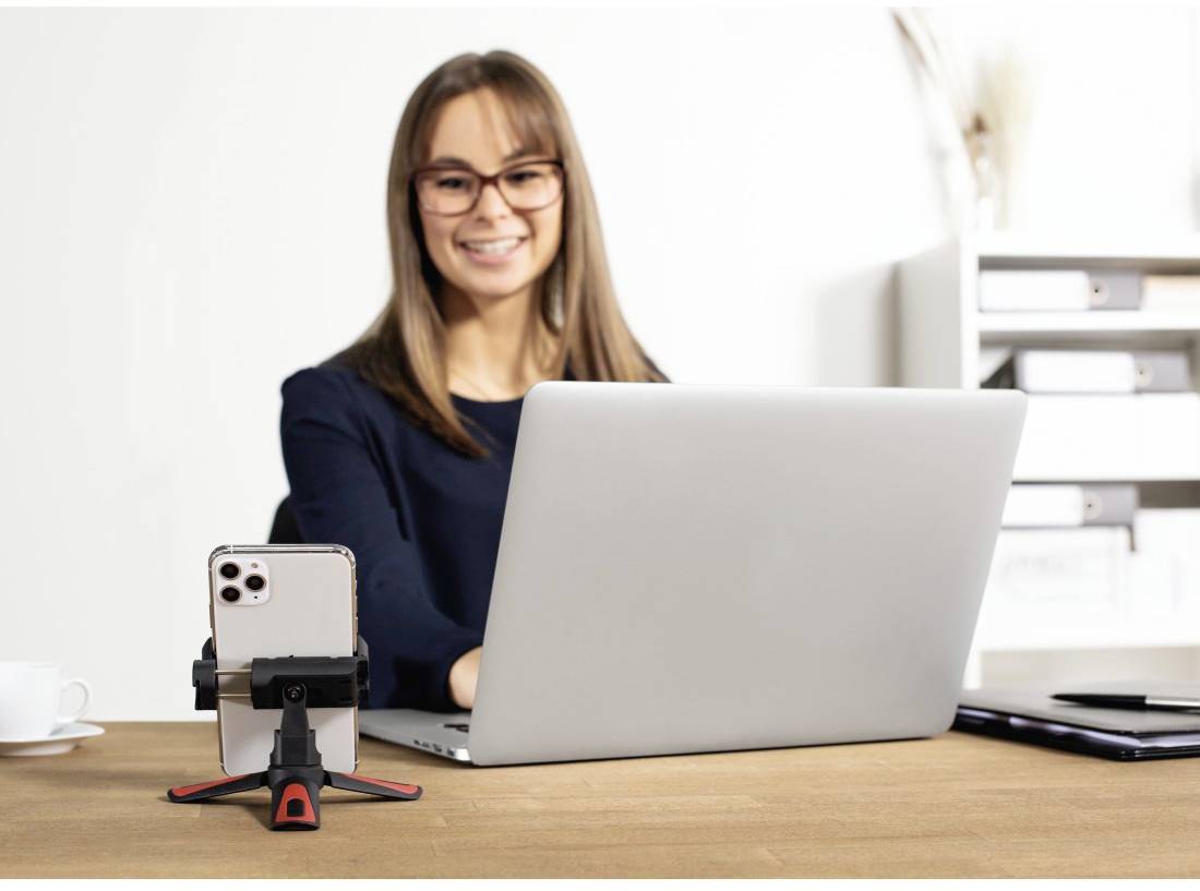 Une femme est assise à son bureau et travaille sur un ordinateur portable. Devant elle, un smartphone est placé sur un trépied. À l'arrière-plan, on aperçoit une étagère.