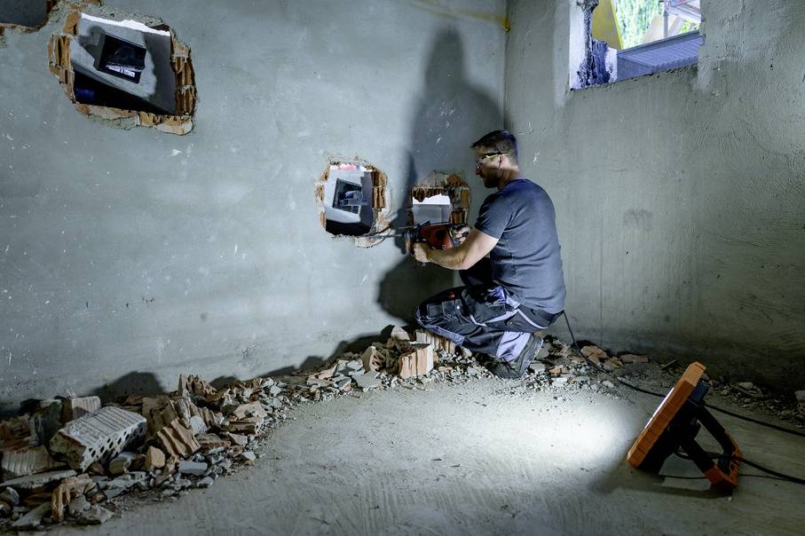 Un artisan travaille dans une pièce inachevée, installant des découpes pour câbles sur un mur. Chantier avec des outils et des débris.