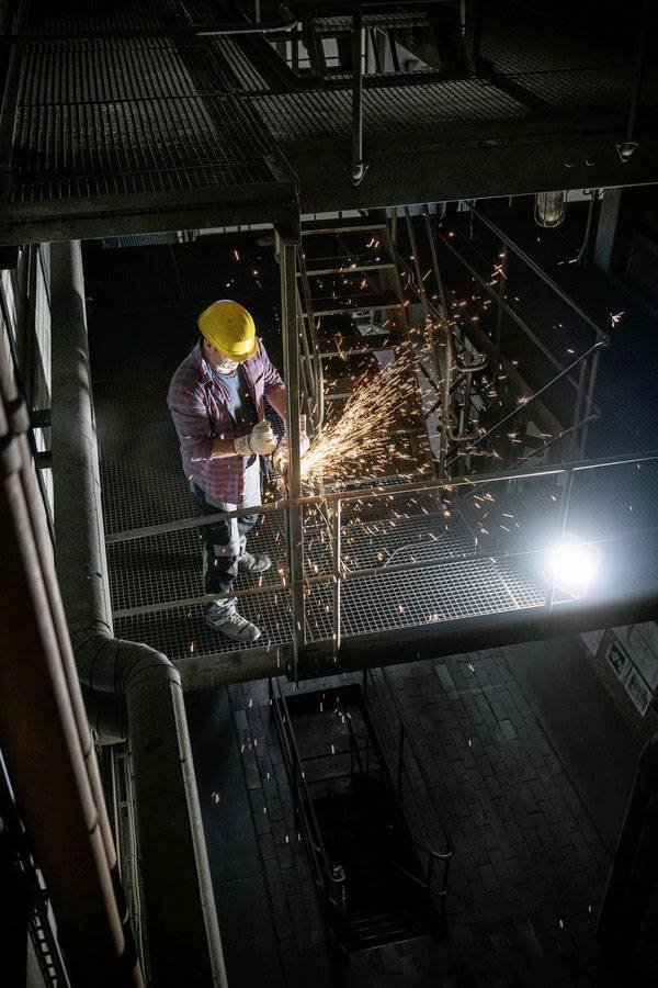 Un ouvrier portant un casque jaune coupe du métal avec une volée d'étincelles dans un environnement industriel. Un échafaudage grillagé est visible aux alentours.