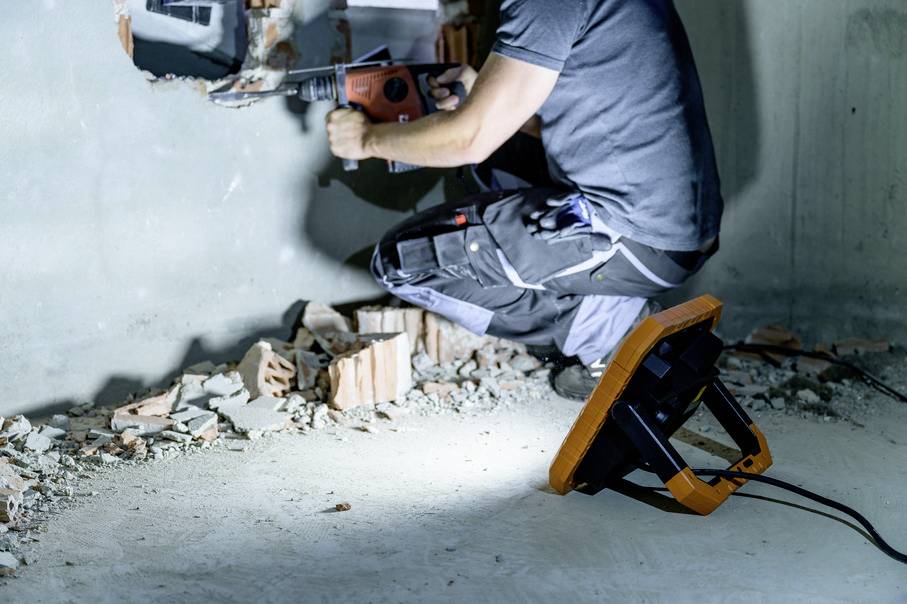 Une personne travaille avec une perceuse sur un mur dans une pièce en cours de rénovation. Des débris et une lampe de travail sont visibles au sol.