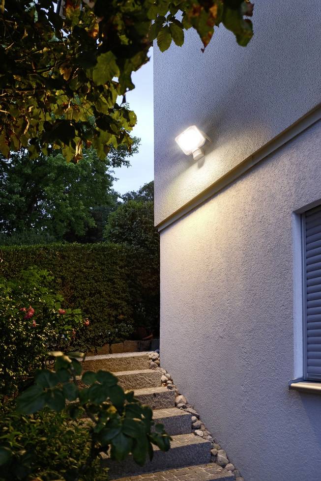 Une lampe extérieure éclaire le soir un escalier situé à côté d'un bâtiment. Des plantes et des arbustes entourent la zone, ajoutant un contraste supplémentaire.
