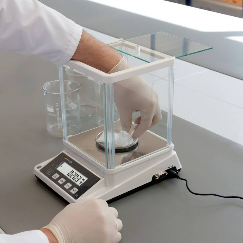 Une personne pèse une substance sur une balance de laboratoire numérique. Un bécher est posé à côté de la balance.