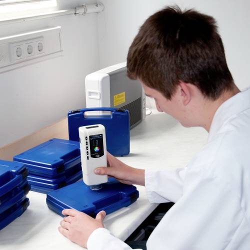 Un homme teste des échantillons bleus à l'aide d'un spectrophotomètre portable sur une table blanche. Des boîtes bleues sont visibles en arrière-plan.