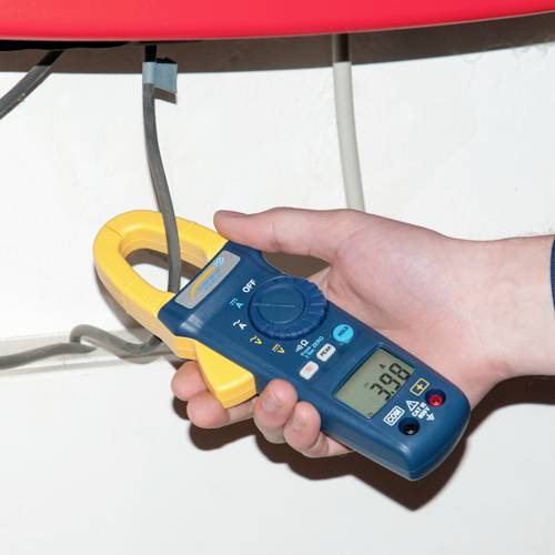 Une main tient un multimètre numérique à pince, mesurant le courant électrique d'un câble gris. L'affichage indique la valeur de 38,8.