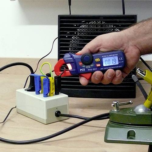 Un artisan mesure le courant électrique dans un câble à l'aide d'une pince ampèremétrique. En arrière-plan, un tableau de distribution électrique.