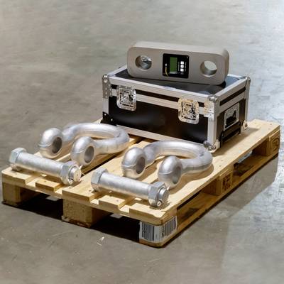 Système de levage de charge lourde sur palette en bois : deux grands manilles métalliques à côté d'un caisson de transport avec un appareil de mesure.