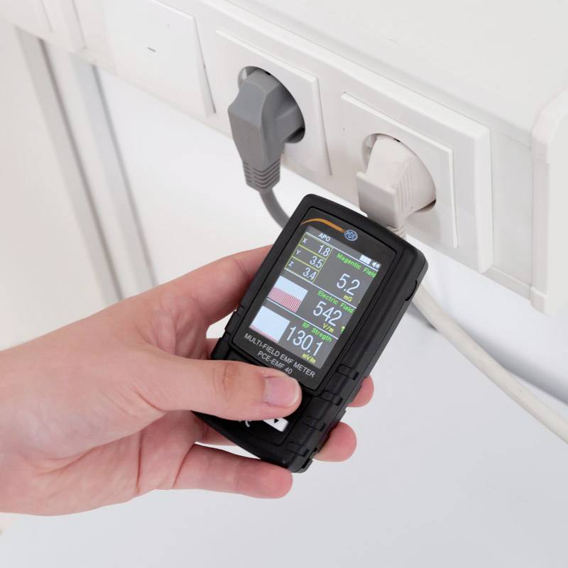 Une main tient un dispositif de surveillance de performance qui mesure des données à partir d'une multiprise. Les affichages montrent les volts, ampères et watts.