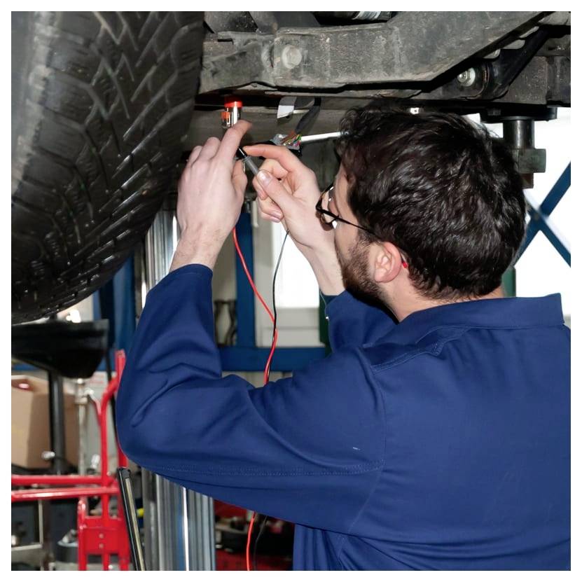 Mécanicien en combinaison bleue utilise un testeur de lumière et une sonde pour inspecter le câblage sous un véhicule sur un pont élévateur.