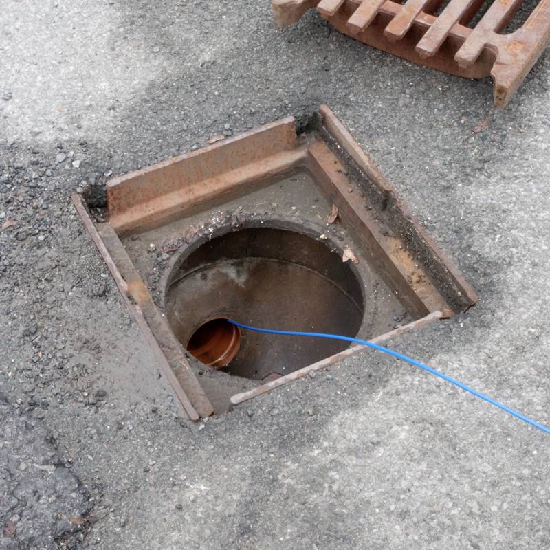 Une bouche d'égout ouverte avec un tuyau d'évacuation visible et un câble bleu à l'intérieur, sur une route asphaltée.