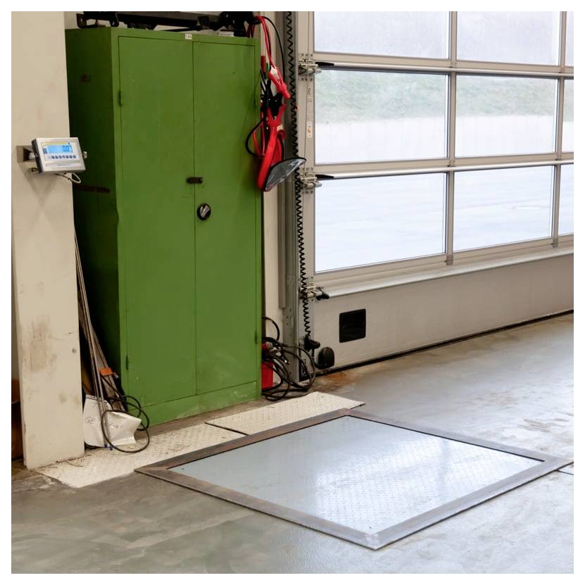 Intérieur de garage industriel avec un grand armoire verte, un panneau de commande fixé au mur, des outils suspendus et une trappe d'accès au sol près de la porte du garage.
