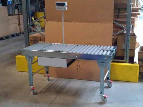 Un chariot de transport surmonté d'une balance, placé dans un environnement d'entrepôt avec des cartons en arrière-plan.