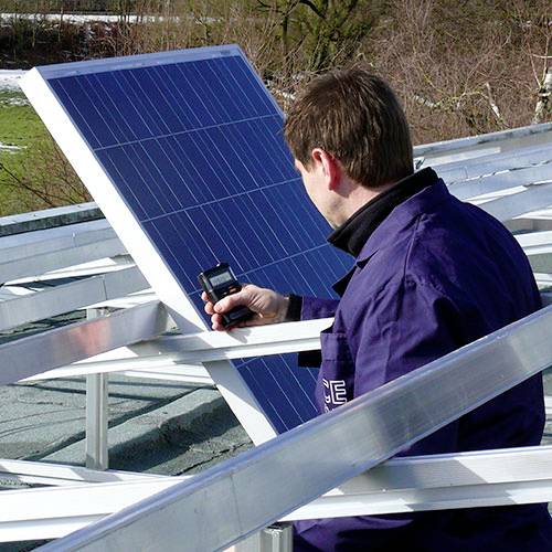 Une personne installe ou vérifie un panneau solaire sur un toit. Elle tient un appareil de mesure. L'arrière-plan montre des arbres et un pré.
