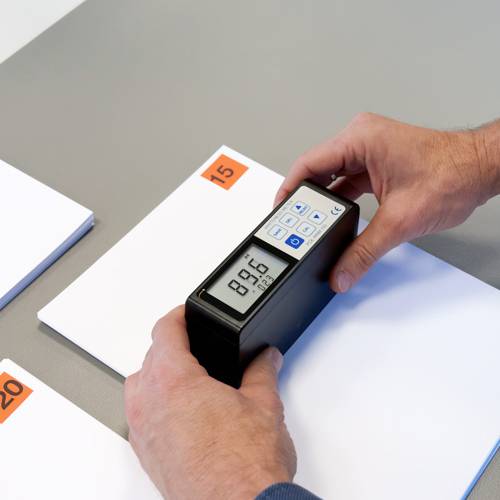 Une personne tient un appareil pour mesurer l'épaisseur d'un objet blanc. L'écran affiche '89,6'. Des cartes numérotées sont visibles en arrière-plan.