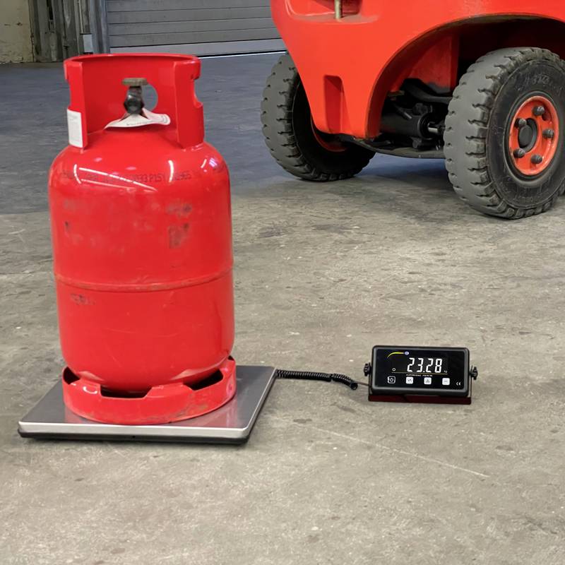 Bouteille de gaz propane rouge posée sur une balance à côté d'un chariot élévateur. L'affichage indique le poids de la bouteille à 23,28 kg.