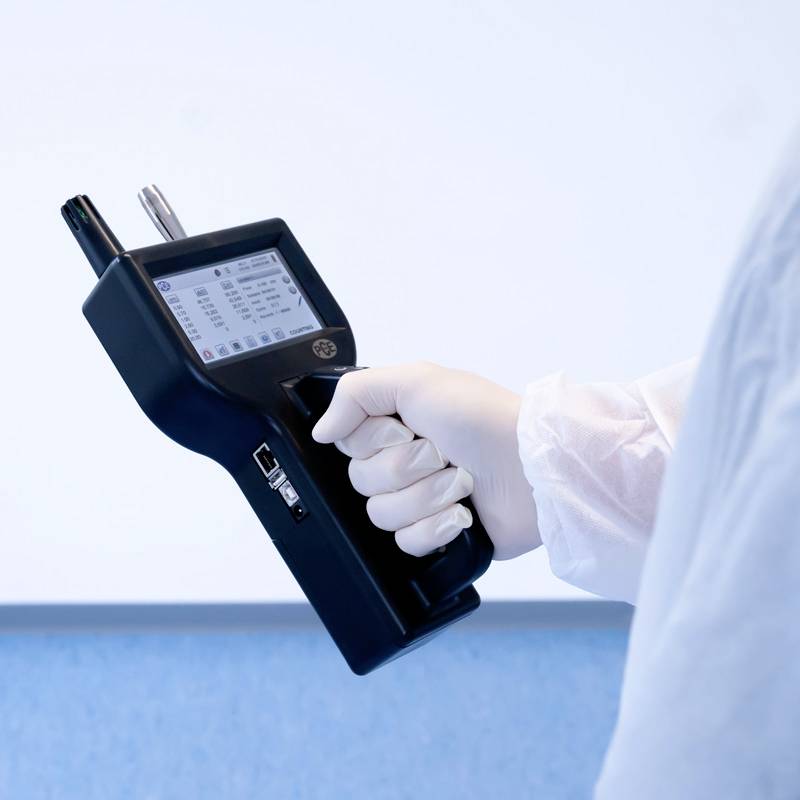 Une personne en tenue de protection tient un appareil portable pour mesurer les particules atmosphériques ; l'accent est mis sur la mesure de précision dans un environnement de laboratoire.