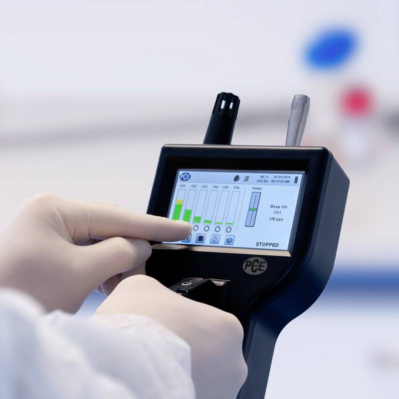 Une main portant une blouse de laboratoire manipule un appareil portable doté d'un écran de mesures. L'écran affiche différentes valeurs et graphiques à barres.