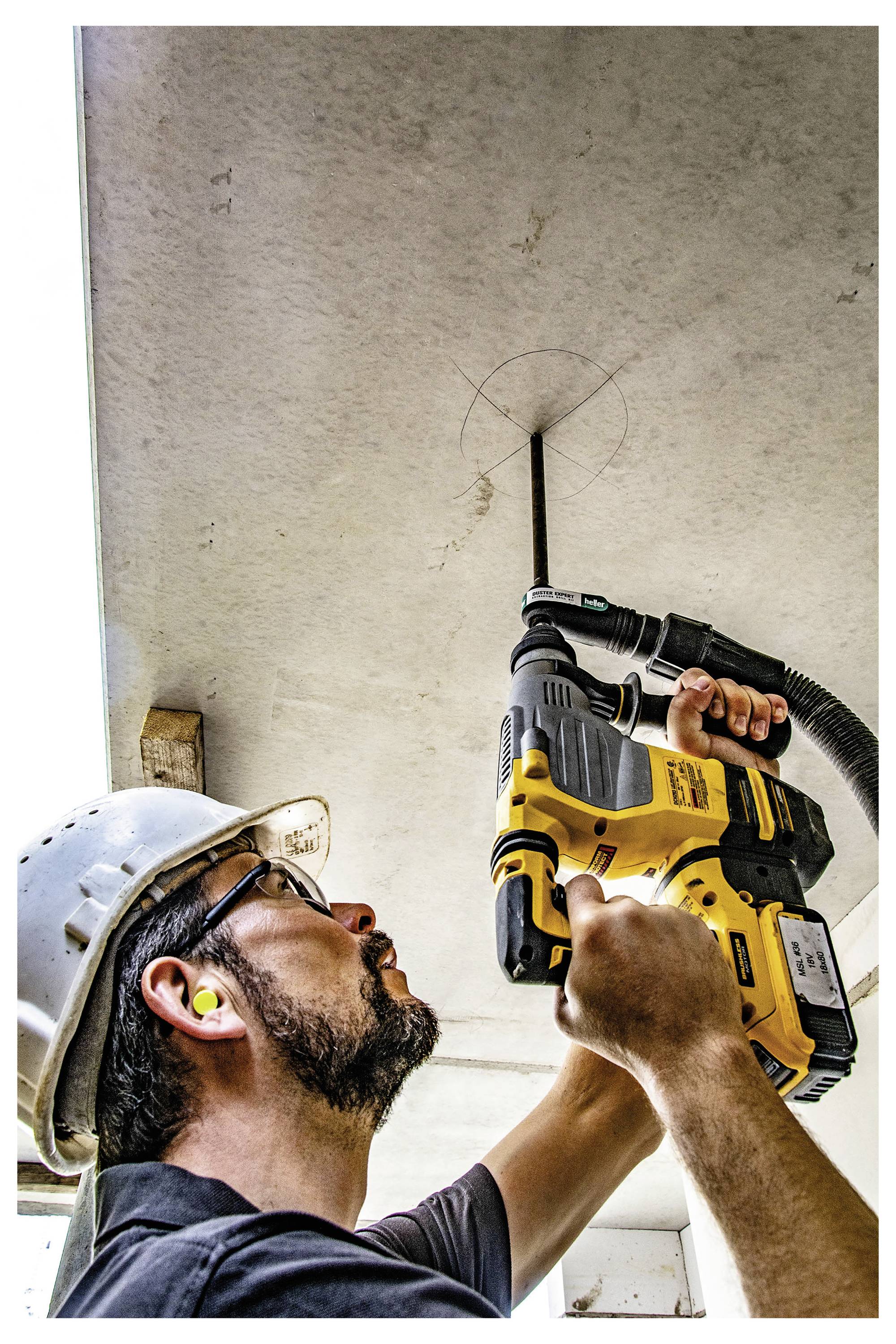 Un ouvrier du bâtiment utilisant une perceuse électrique jaune au-dessus de sa tête pour percer un plafond, portant un casque de protection et une protection auditive.