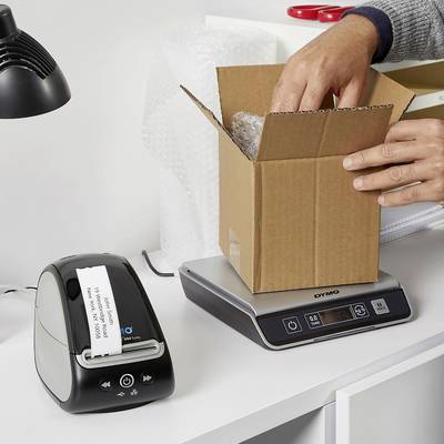 Une personne emballe un petit objet dans un carton sur une table avec une balance électronique et une imprimante d'étiquettes.