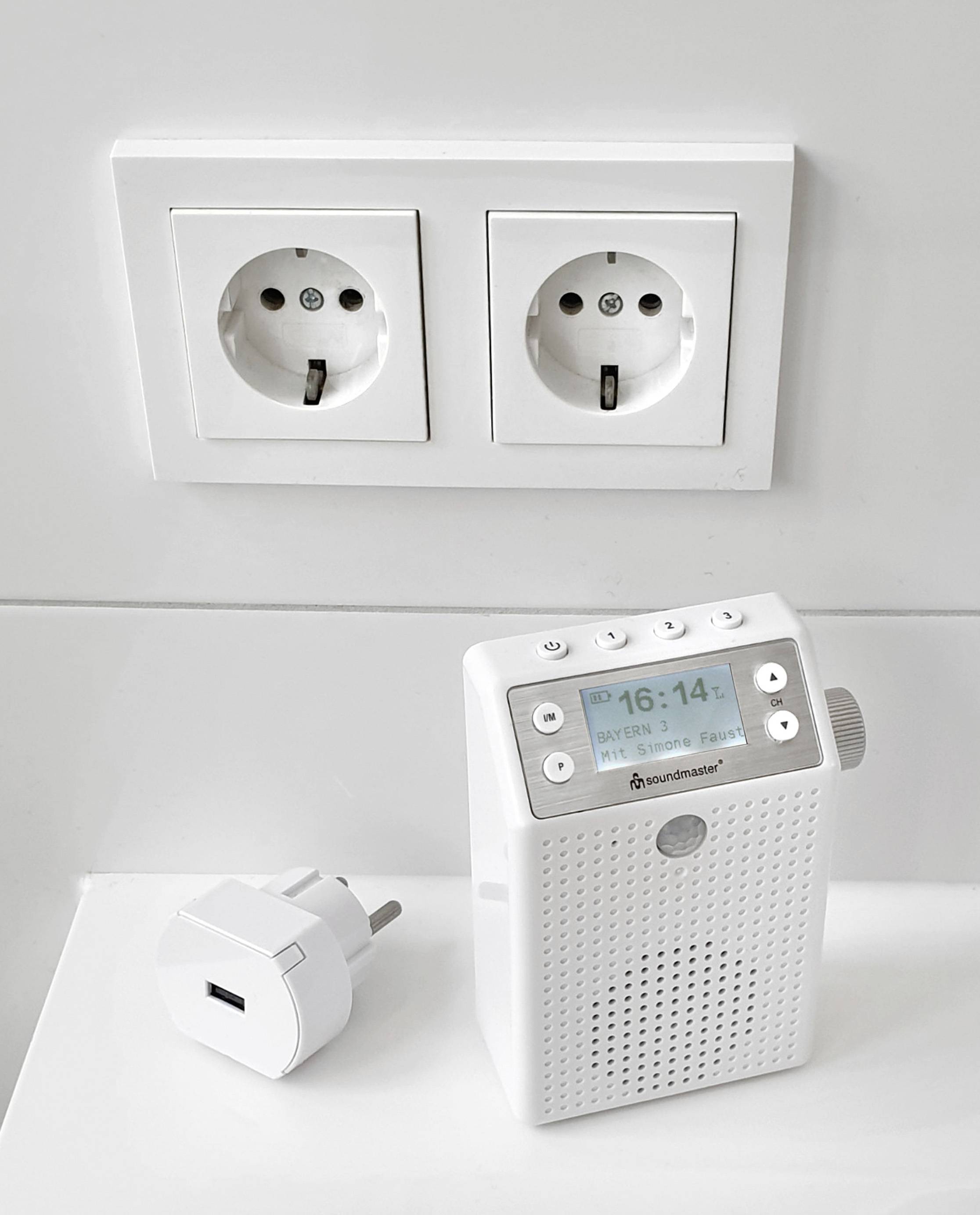 Un poste de radio FM blanc avec un affichage numérique indique '16:14'. À côté, un adaptateur est branché sur deux prises doubles contre un mur blanc.