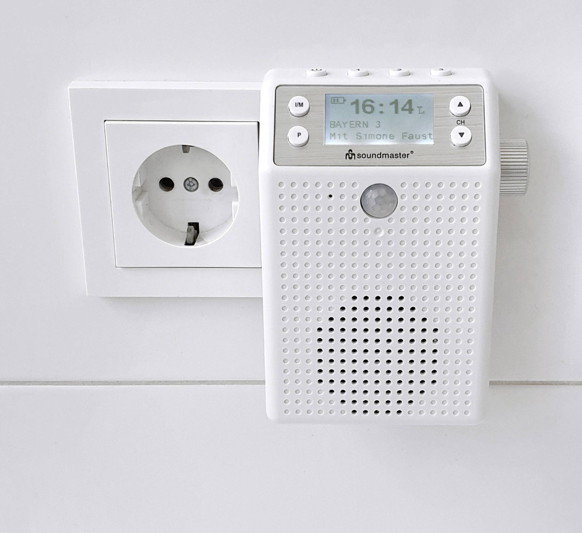 Un poste de radio blanc, branché à une prise électrique, affiche l'heure 16:14 et la station de radio Bayern 3 sur son écran.