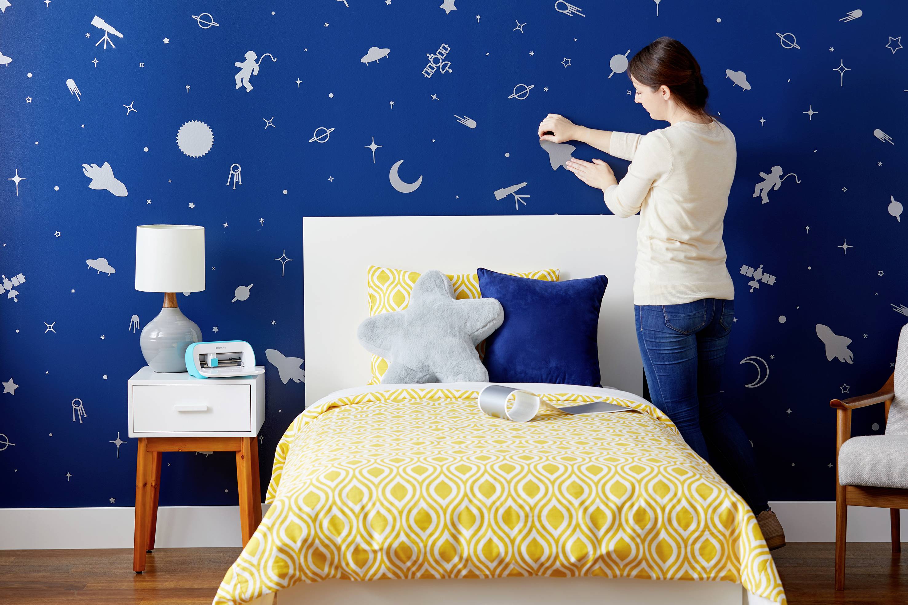 Une femme décore un mur de chambre d'enfant avec des motifs d'espace. Le lit est orné d'un couvre-lit jaune à motifs et de coussins en forme d'étoiles.