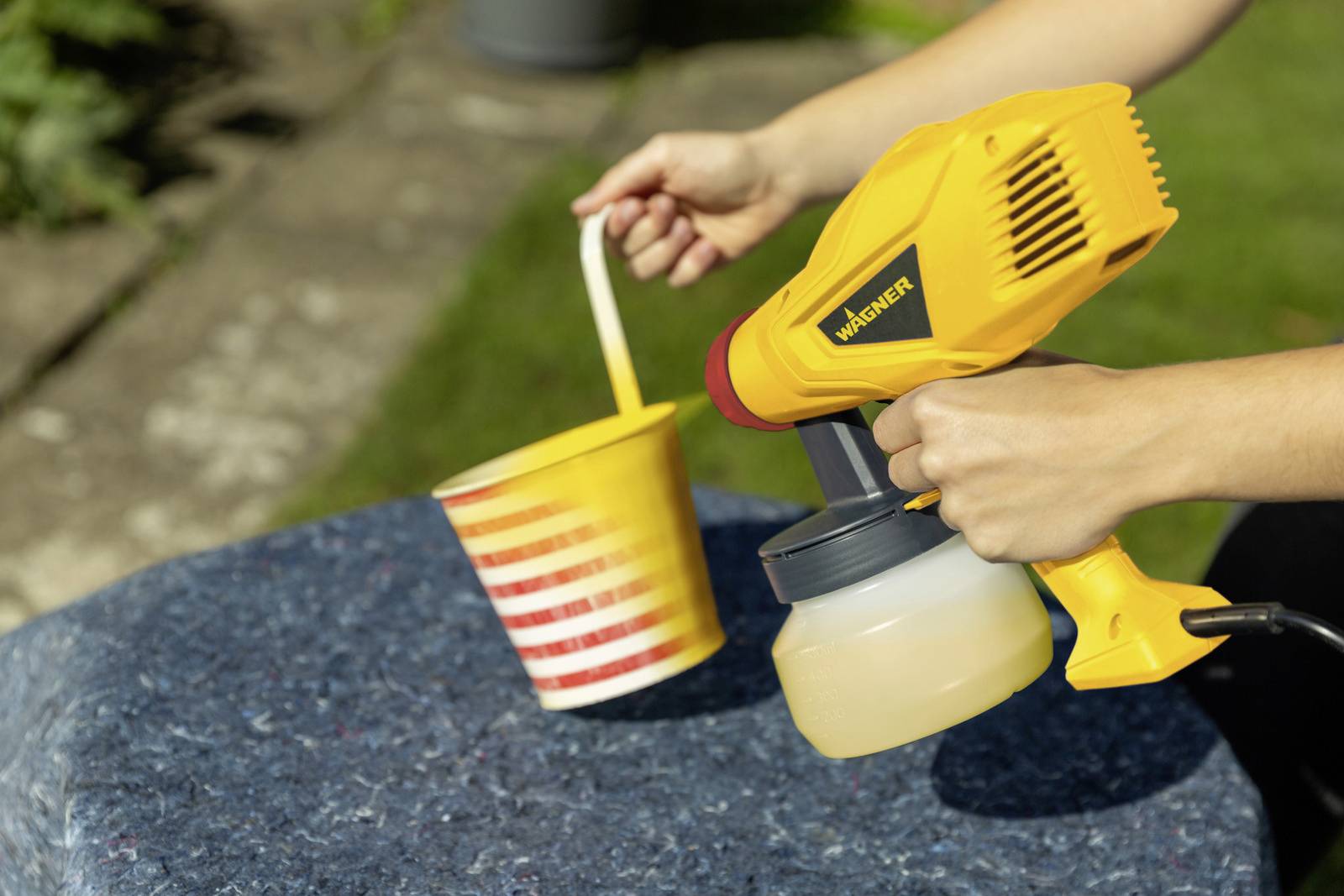 Une personne tient un pistolet à eau jaune et peint une tasse rayée jaune et rouge sur une table bleue à l'extérieur.