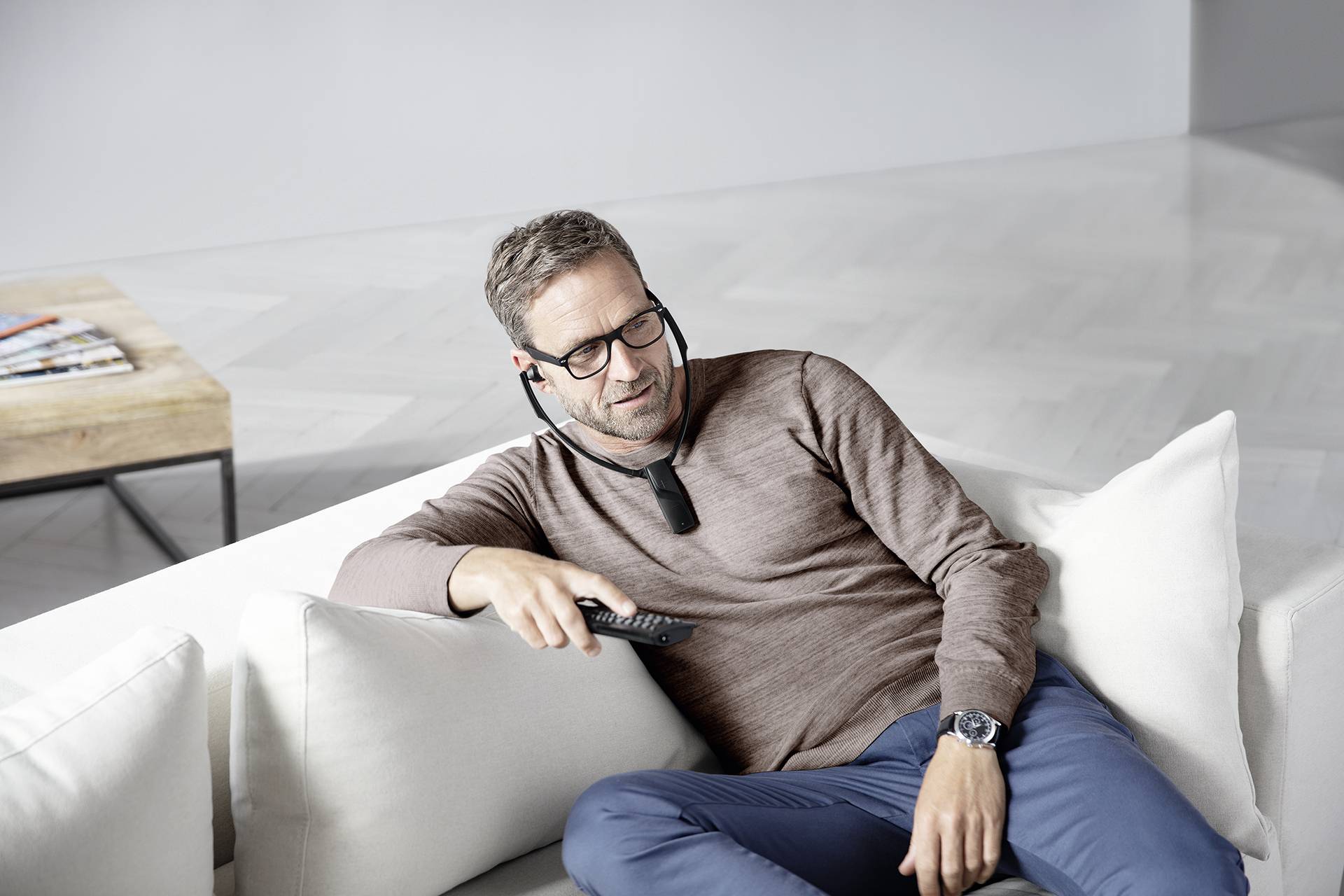 Un homme portant des lunettes et des écouteurs est allongé de manière détendue sur un canapé et tient une télécommande à la main, avec une table portant des magazines en arrière-plan.