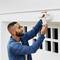 Un homme portant une chemise en jean installe une caméra extérieure blanche sur le mur d'une maison, vue de dessous, l'accent étant mis sur le dispositif de sécurité.