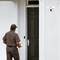 Un livreur se tient devant une porte d'entrée, tenant un colis dans sa main. À côté de la porte est installée une caméra de sécurité moderne.