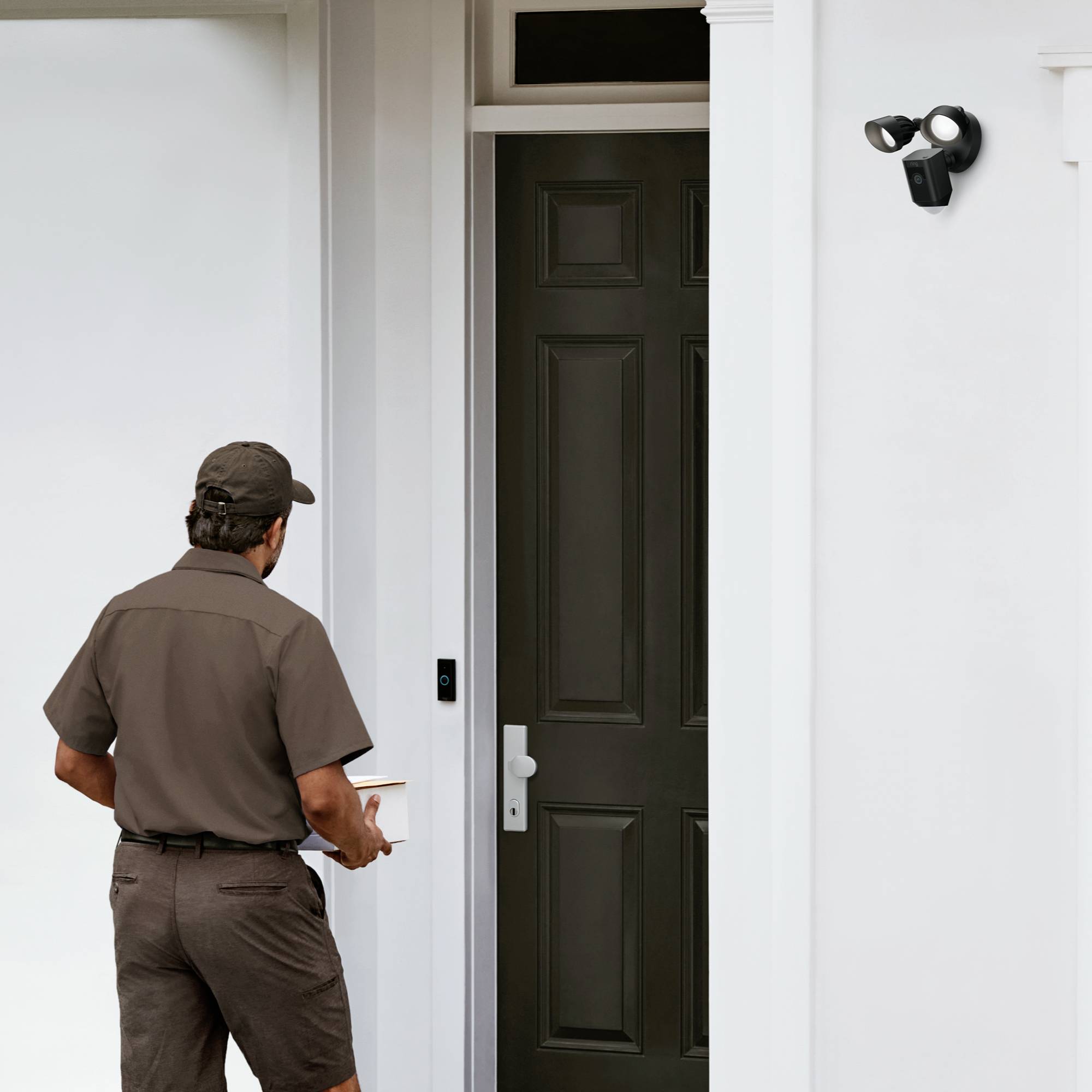 Un facteur se tient devant une porte d'entrée et tient un colis dans sa main. Au-dessus de la porte, une caméra de sécurité est installée.