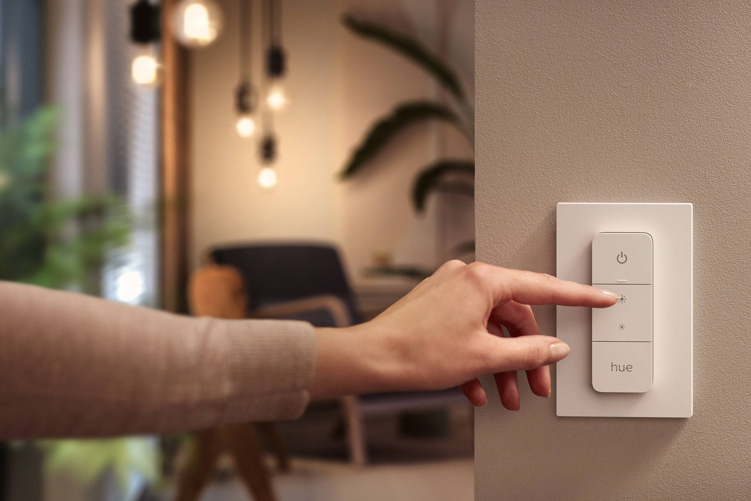 Une main appuie sur un interrupteur portant l'inscription 'hue' dans une pièce aux lumières chaleureuses, avec des lampes suspendues en arrière-plan.